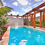 Modern water pool closes up to the blue water with a garden, poolside with wooden pillars and beams under blue sky and white clouds