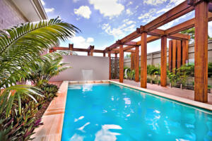 Modern water pool closes up to the blue water with a garden, poolside with wooden pillars and beams under blue sky and white clouds