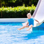 Happy laughing little boy playing on water slide in outdoor swimming pool on a hot summer day. Kids learn to swim. Child wearing sun protection rash guard sliding on aqua playground in tropical resort