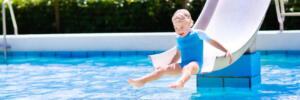 Happy laughing little boy playing on water slide in outdoor swimming pool on a hot summer day. Kids learn to swim. Child wearing sun protection rash guard sliding on aqua playground in tropical resort
