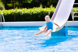 Happy laughing little boy playing on water slide in outdoor swimming pool on a hot summer day. Kids learn to swim. Child wearing sun protection rash guard sliding on aqua playground in tropical resort